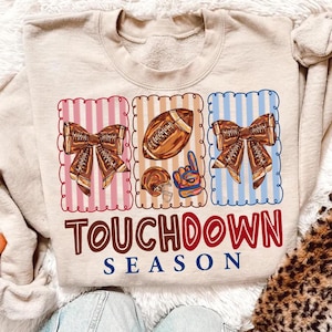 May include: Beige sweatshirt with a football-themed design. The design includes a football, a helmet, a bow, and the words "Touchdown Season" in red and blue. The background has pink, beige, and blue stripes.
