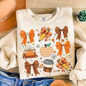 Puede incluir: Una sudadera beige con un diseño de temática otoñal que presenta una pila de libros, una manzana a cuadros, una taza de café, lápices y lazos en naranja, marrón y blanco.