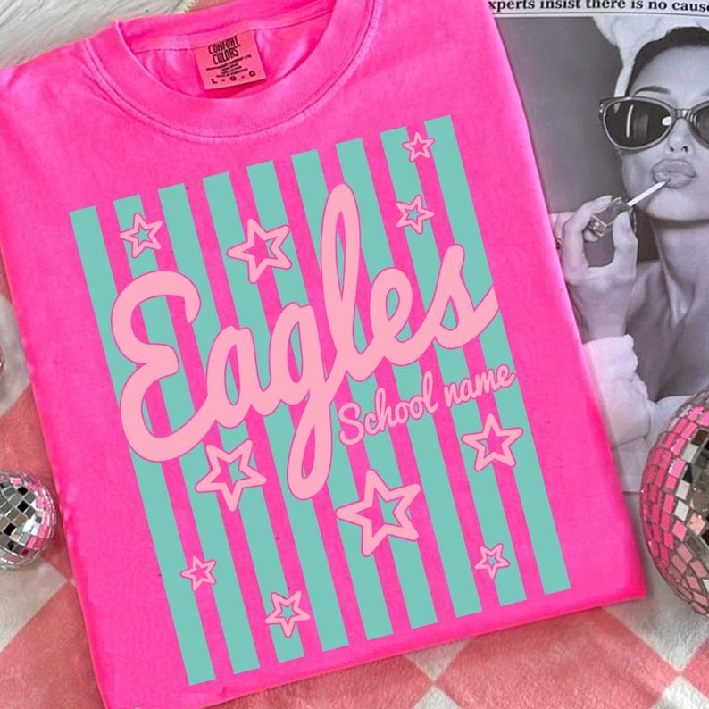 Puede incluir: Camiseta rosa brillante con la palabra "Eagles" en cursiva, acentuada por rayas y estrellas turquesas. Las palabras "School name" est&aacute;n debajo de la palabra "Eagles". La camiseta est&aacute; hecha de un material suave.