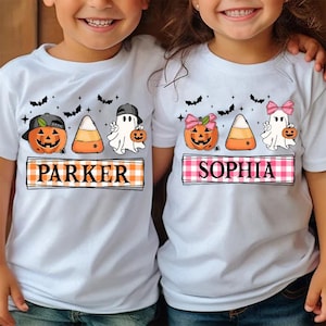 以下が含まれることがあります： ハロウィンをテーマにした白いTシャツ2枚。シャツには、カボチャ、コーン、ゴーストが描かれており、下に「Parker」と「Sophia」の名前がプリントされています。