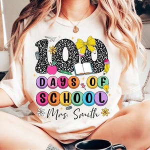 May include: A white t-shirt with a black and white graphic of the number 100 with school supplies. The text "100 Days of School" is in colourful circles. The text "Mrs. Smith" is below the graphic.