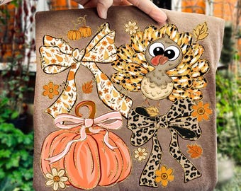 May include: Brown t-shirt featuring a fall-themed design. The design includes a cartoon turkey, pumpkins, bows, and floral accents in shades of orange, brown, and gold. The turkey has large eyes and a red tongue. The bows have floral and leopard prints.