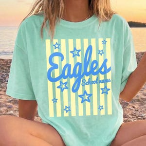 Puede incluir: Una camiseta verde claro con la palabra "Eagles" en escritura azul, rodeada de estrellas azules. La palabra "Eagles" est&aacute; sobre un fondo a rayas amarillas. Las palabras "School name" est&aacute;n debajo de la palabra "Eagles".