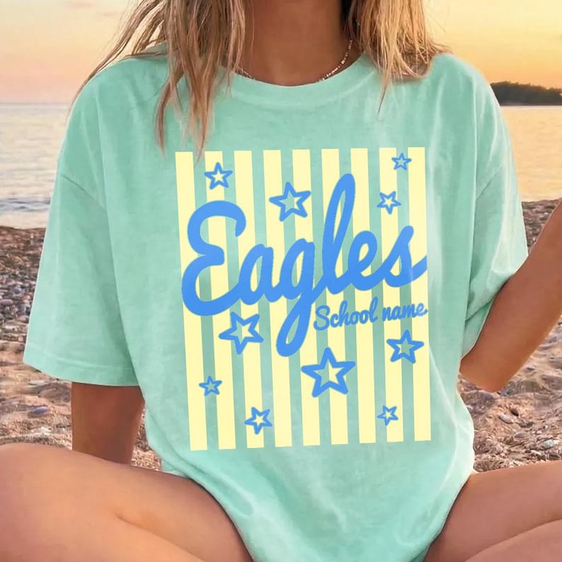 Puede incluir: Una camiseta verde claro con la palabra "Eagles" en escritura azul, rodeada de estrellas azules. La palabra "Eagles" est&aacute; sobre un fondo a rayas amarillas. Las palabras "School name" est&aacute;n debajo de la palabra "Eagles".