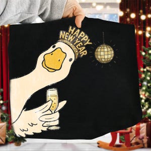 May include: Black graphic tee featuring a cartoon goose wearing a crown with "HAPPY NEW YEAR" text. The goose holds a champagne glass and a disco ball is nearby. Festive design for New Year's celebrations.