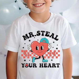 May include: White t-shirt with a red heart graphic wearing a blue hat and holding peace signs. The heart has a smiling face and the text "MR. STEAL YOUR HEART" is printed in black.
