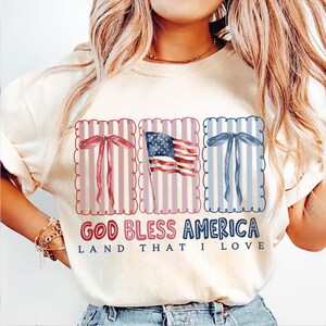 May include: Cream-colored sweatshirt with a patriotic design. The graphic includes a US flag and decorative bows in red and blue, with the text "GOD BLESS AMERICA LAND THAT I LOVE".