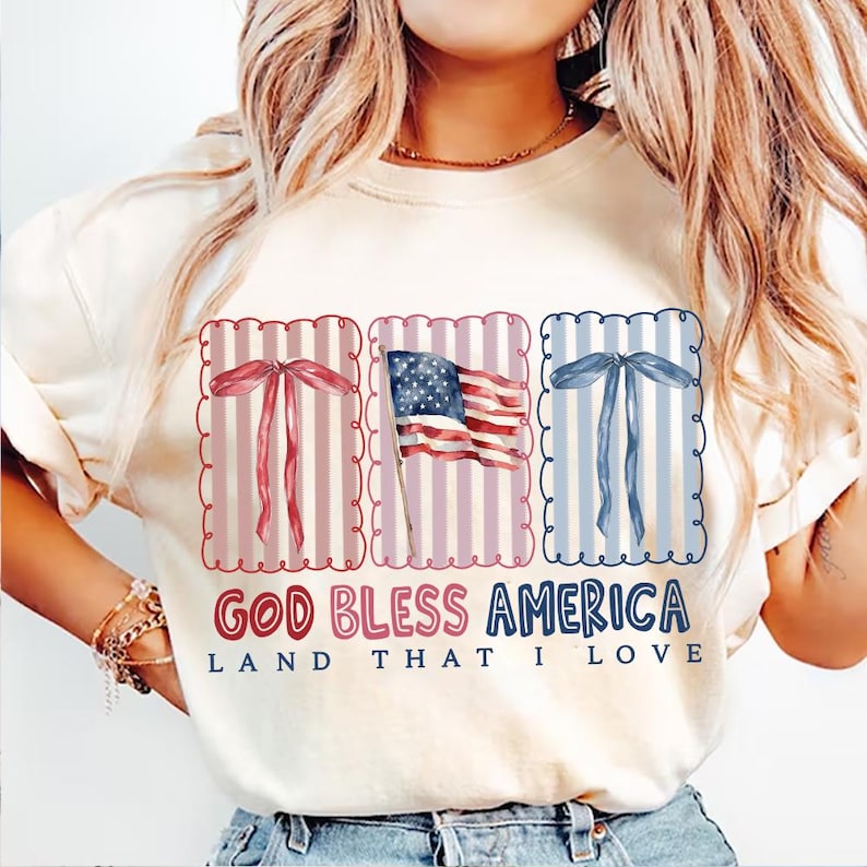 May include: Cream-colored sweatshirt with a patriotic design. The graphic includes a US flag and decorative bows in red and blue, with the text "GOD BLESS AMERICA LAND THAT I LOVE".
