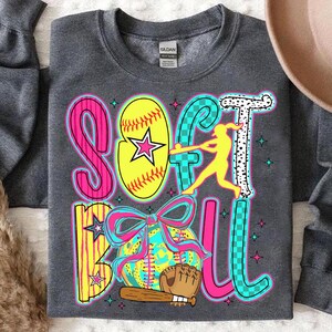 May include: A gray sweatshirt with the word "Softball" written in colorful letters. The letters are decorated with stars, checkered patterns, and a softball player silhouette. A softball with a bow and a baseball bat are also included in the design.