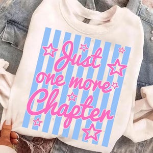 May include: White sweatshirt with a blue and white striped design. The text "Just one more Chapter" is in pink script with pink star accents. The sweatshirt is made of a soft material.