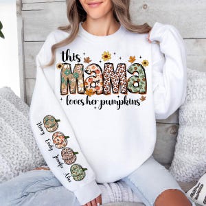 Puede incluir: Sudadera blanca con el texto "this mama loves her pumpkins" en letras de colores con detalles florales y de estampado de leopardo. La manga tiene una fila vertical de cuatro calabazas con los nombres "Henry, Emily, Jennifer, Alice" escritos a su lado.