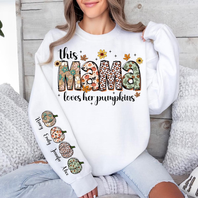 Puede incluir: Sudadera blanca con el texto "this mama loves her pumpkins" en letras de colores con detalles florales y de estampado de leopardo. La manga tiene una fila vertical de cuatro calabazas con los nombres "Henry, Emily, Jennifer, Alice" escritos a su lado.
