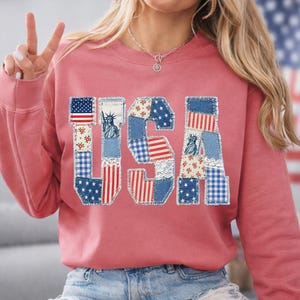 K&ouml;nnte beinhalten: Ein rosafarbenes Sweatshirt mit dem Wort "USA" auf der Vorderseite. Die Buchstaben bestehen aus Patchwork-Denim und Stoff in rot, wei&szlig; und blau, einschlie&szlig;lich Sternen, Streifen und der Freiheitsstatue.
