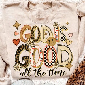 May include: Beige sweatshirt with the phrase "GOD IS GOOD all the time" in a colorful, retro design. The text incorporates leopard print, cherries, and a smiley face. Decorative hearts and stars surround the text.