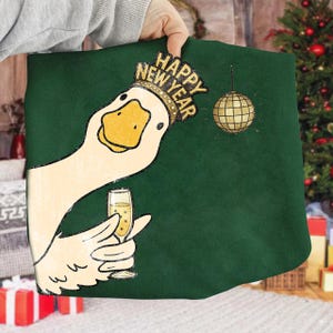 May include: A green fabric item featuring a cartoon goose wearing a "Happy New Year" crown and holding a champagne glass. A gold disco ball hangs in the background. The goose has a yellow beak and is set against a green background.