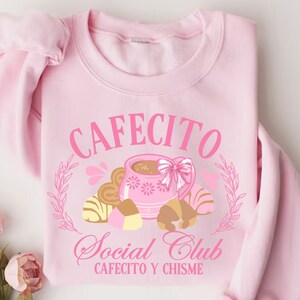May include: Pink sweatshirt with a graphic design of a coffee cup and pastries. The text reads "Cafecito Social Club Cafecito y Chisme".