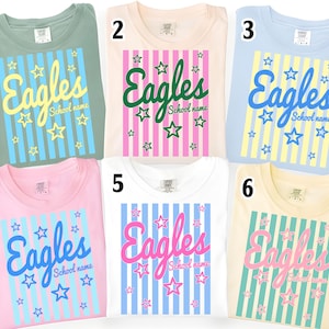Puede incluir: Seis camisetas de diferentes colores, cada una con la palabra "Eagles" en escritura cursiva, con el nombre de la escuela debajo. El dise&ntilde;o incluye estrellas y rayas verticales.