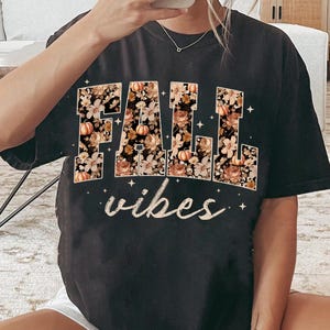 May include: Black t-shirt with the words "FALL vibes" in a floral design with pumpkins. The letters are filled with a pattern of flowers and pumpkins in shades of orange, brown, and cream. The word "vibes" is in a cursive font.