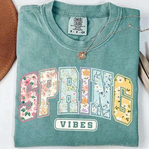 Puede incluir: Una camiseta Comfort Colors de color turquesa con la palabra "SPRING" en un dise&ntilde;o floral y la palabra "VIBES" debajo. Un collar dorado con un colgante est&aacute; sobre la camisa.