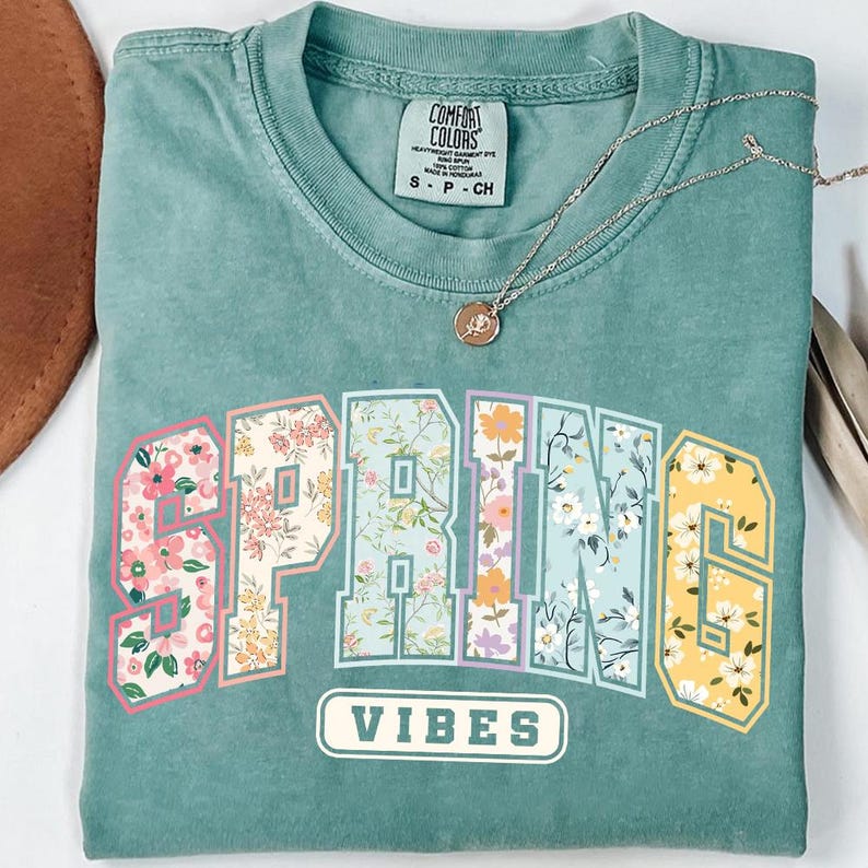 Puede incluir: Una camiseta Comfort Colors de color turquesa con la palabra "SPRING" en un dise&ntilde;o floral y la palabra "VIBES" debajo. Un collar dorado con un colgante est&aacute; sobre la camisa.