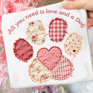 May include: White fabric with a patchwork paw print design. The paw print is made of various pink and cream fabrics, including floral, polka dot, and gingham patterns. The text "All you need is love and a Dog" is embroidered in pink.