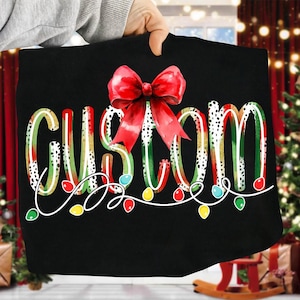 May include: Black t-shirt with the word "CUSTOM" in a festive design. The letters are outlined in white, with green and red stripes, and black and white polka dots. A red bow is at the top, and a string of colorful lights is at the bottom.