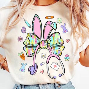 May include: Cream-colored t-shirt featuring a colorful Easter-themed design. The design includes a stethoscope, bunny ears, a bow, and various Easter-related graphics like eggs, carrots, and flowers. The design is in pastel colors with gold accents.