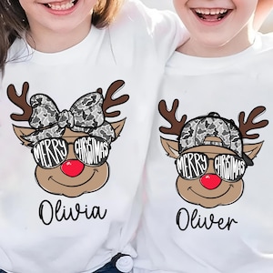 Può includere: Magliette bianche con disegni di renne natalizie abbinate. Una renna ha un fiocco, l'altra un cappello, entrambi con motivi mimetici. Ogni renna indossa occhiali da sole con "Merry Christmas" e ha un naso rosso. I nomi "Olivia" e "Oliver" sono sotto.