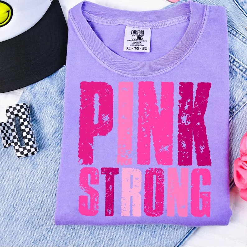 May include: A lilac Comfort Colors t-shirt with the words "PINK STRONG" in shades of pink. The shirt is folded on a denim surface, with a black and white checkered accessory and a black and white hat in the background.