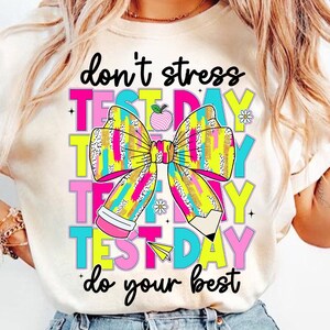May include: White t-shirt with a colorful graphic that says "don't stress test day do your best". The graphic features a large bow with a pencil and an apple.