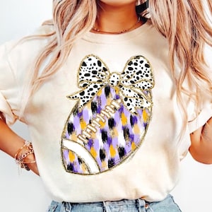 May include: A white t-shirt with a purple, black, and gold football design. The football is decorated with a black and white polka dot bow.