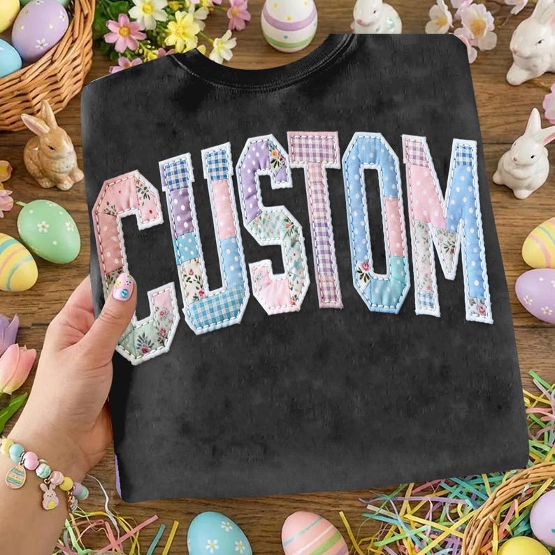 May include: Black sweatshirt with the word "CUSTOM" appliqued across the front in a patchwork design. The letters are made of various pastel-colored fabrics, including floral and gingham patterns. The sweatshirt is surrounded by Easter decorations.