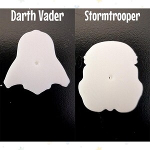 Op de afbeelding: Twee witte, gestileerde karakteruitsparingen, &eacute;&eacute;n die op Darth Vader lijkt en de andere op een Stormtrooper-helm. De tekst "Darth Vader" en "Stormtrooper" staan boven elke uitsparing. De uitsparingen staan op een zwarte achtergrond.