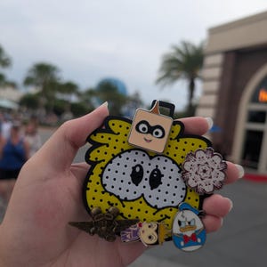May include: A collection of colorful enamel pins featuring cartoon characters and designs. The pins include a yellow and black character with white polka dots, a square-shaped character, a snowflake, and Donald Duck.