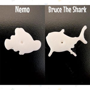 Op de afbeelding: Twee witte plastic uitsparingen, &eacute;&eacute;n in de vorm van Nemo met de tekst "Nemo" erboven, en de andere in de vorm van een haai met de tekst "Bruce The Shark" erboven. Beide hebben een klein gaatje in het midden.