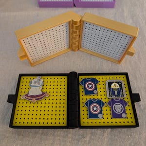 May include: A yellow display case with a black frame, holding various enamel pins. The pins include a pug on a pillow, Captain America shirts, a Star Wars droid, and a Black Panther shirt. The case is open, revealing the pins.