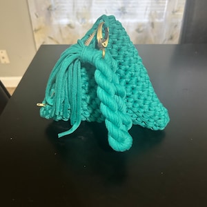 May include: A teal crocheted pouch with a gold zipper and a tassel. The pouch is made of a thick, textured yarn and has a rope-like handle.