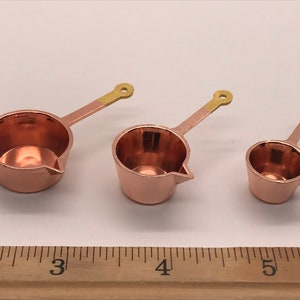 May include: Three copper measuring cups with gold handles. The cups are nested together and measure 5, 7.5, and 10 cm in diameter.
