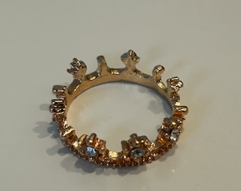 May include: A gold-toned ring with a crown design. The ring is decorated with small, clear rhinestones.
