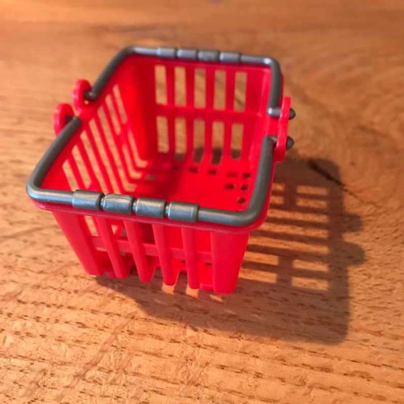 Op de afbeelding: Een klein rood plastic winkelmandje met een grijze metalen rand. Het mandje is leeg en heeft aan elke kant een handvat.