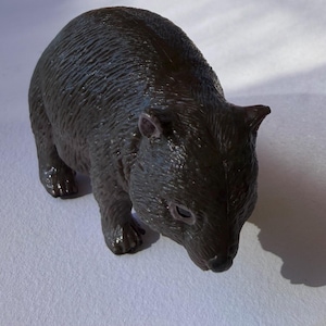 May include: A dark brown wombat figurine. The toy wombat is in a walking position, with visible ears, eyes, and paws. The figurine has a textured surface, and is casting a shadow on the white surface.