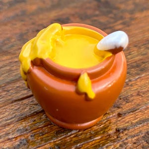 May include: A small, brown ceramic pot filled with yellow honey. A white spoon is partially submerged in the honey.