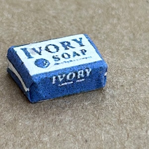 May include: A small blue bar of Ivory soap wrapped in white paper. The bar has the words "Ivory Soap" printed on the front and "Ivory" printed on the bottom.