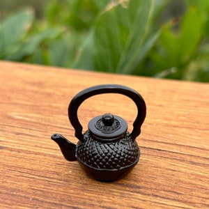 May include: A small black metal teapot with a textured design and a lid. The teapot is sitting on a wooden surface.