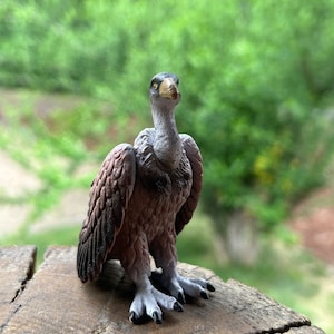 May include: A detailed figurine of a vulture with a grey and brown feathered body, a black beak, and yellow eyes. The figurine is perched on a wooden surface.
