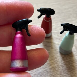 May include: A close-up of three miniature spray bottles. One is held in a person's fingers. The bottles are pink, red, and light green, each with a black spray nozzle and a silver band near the base. The bottles are on a light-colored surface.