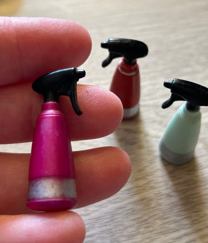 May include: A close-up of three miniature spray bottles. One is held in a person's fingers. The bottles are pink, red, and light green, each with a black spray nozzle and a silver band near the base. The bottles are on a light-colored surface.