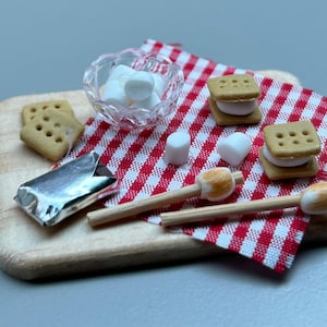May include: A miniature camping scene with a wooden chopping board, a red and white gingham tablecloth, a bowl of marshmallows, graham crackers, chocolate bars, and skewers with marshmallows.