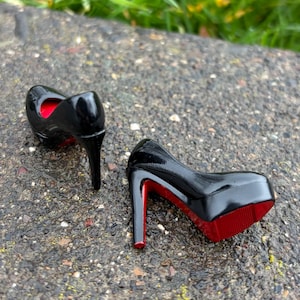 May include: A pair of black high-heeled shoes with red soles and interior. The glossy shoes have a platform sole and a stiletto heel. The shoes are displayed on a textured, gray surface.
