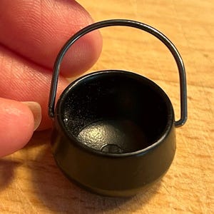 May include: A small black cauldron with a metal handle. The cauldron is about 2 inches tall and 2 inches wide.